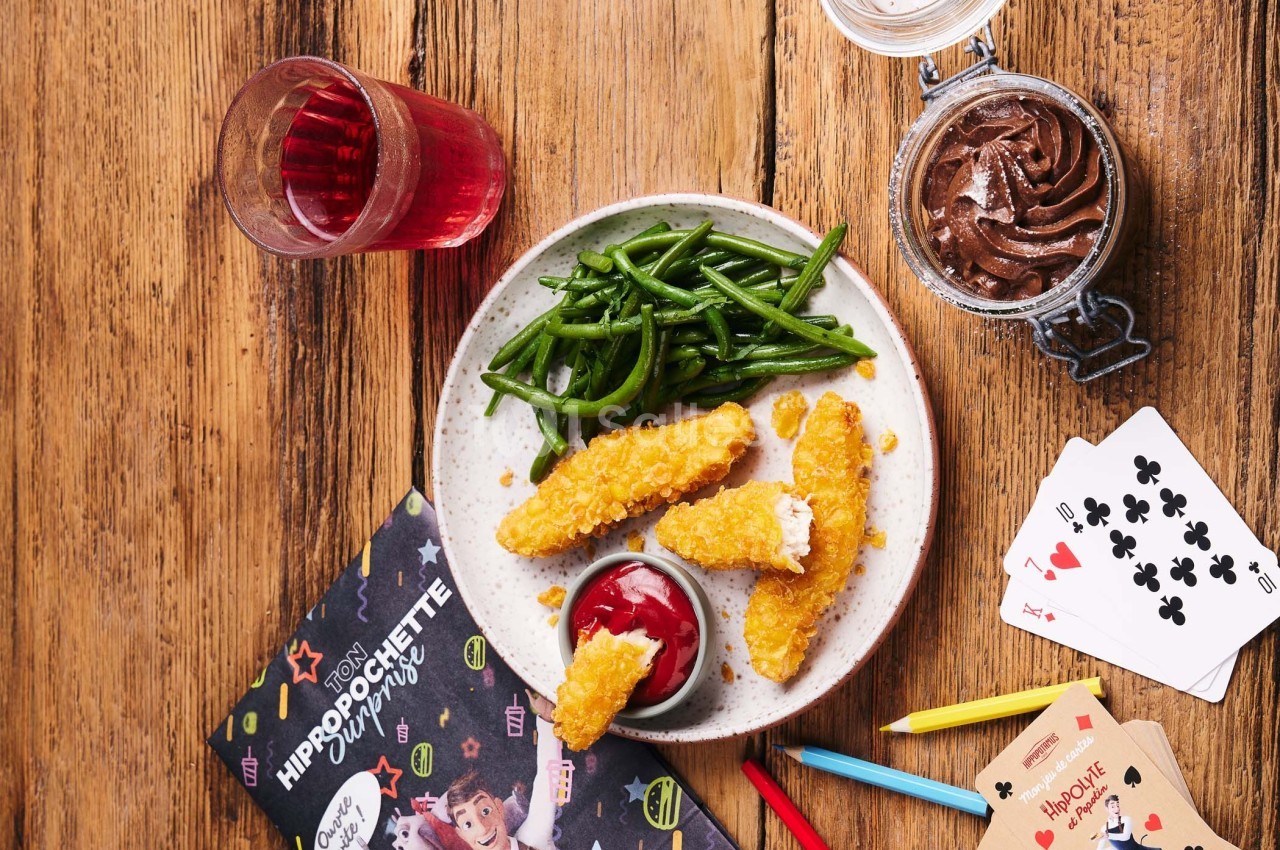 Assiette avec bâtonnets de poulet panés, haricots verts, sauce ketchup, verre de boisson rouge et mousse au chocolat.