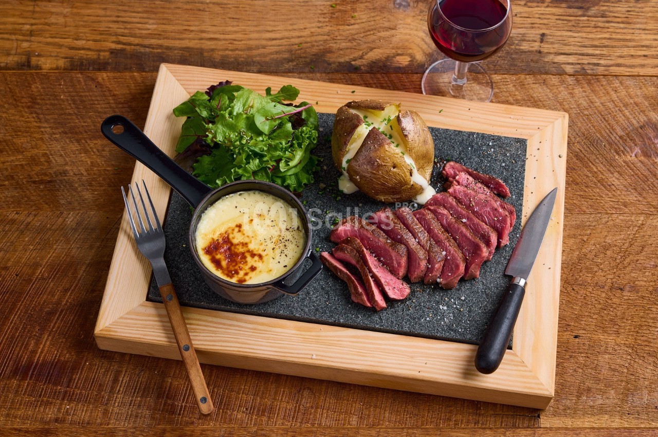 Assiette avec viande tranchée, pomme de terre au four, salade verte et fromage fondu, servie sur une planche en bois.