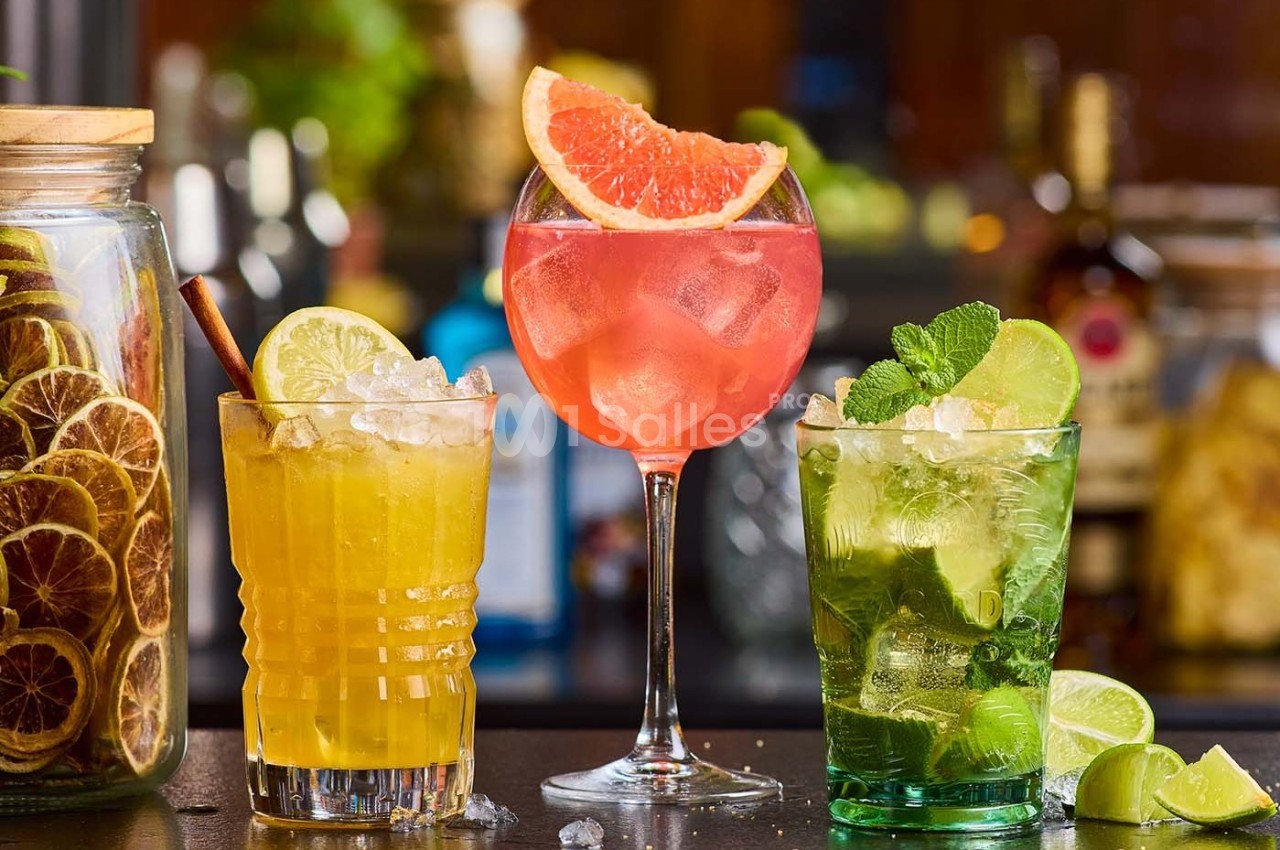 Trois cocktails colorés avec glaçons, garnis de fruits et herbes, posés sur un bar avec un arrière-plan flou.