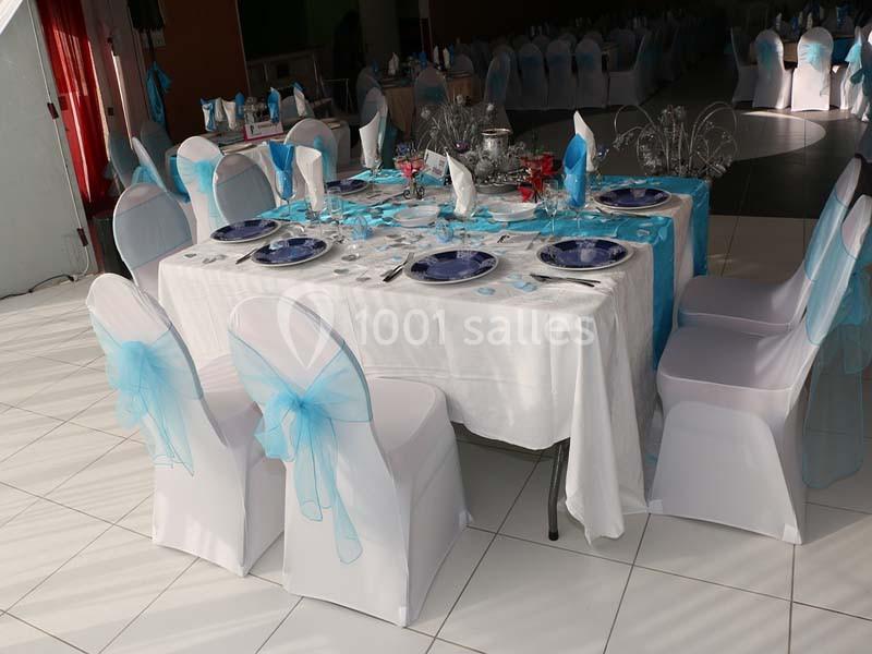 Table élégamment dressée avec nappes blanches, vaisselle bleue et chaises décorées de nœuds en tissu bleu clair.