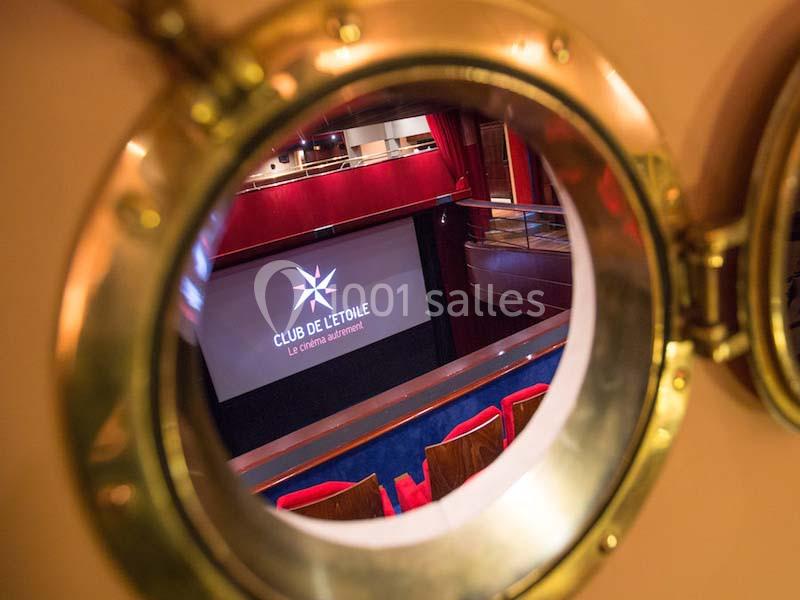 Vue d'une salle de cinéma à travers un hublot, avec un écran affichant ’Club de l'Étoile’ et des sièges rouges.