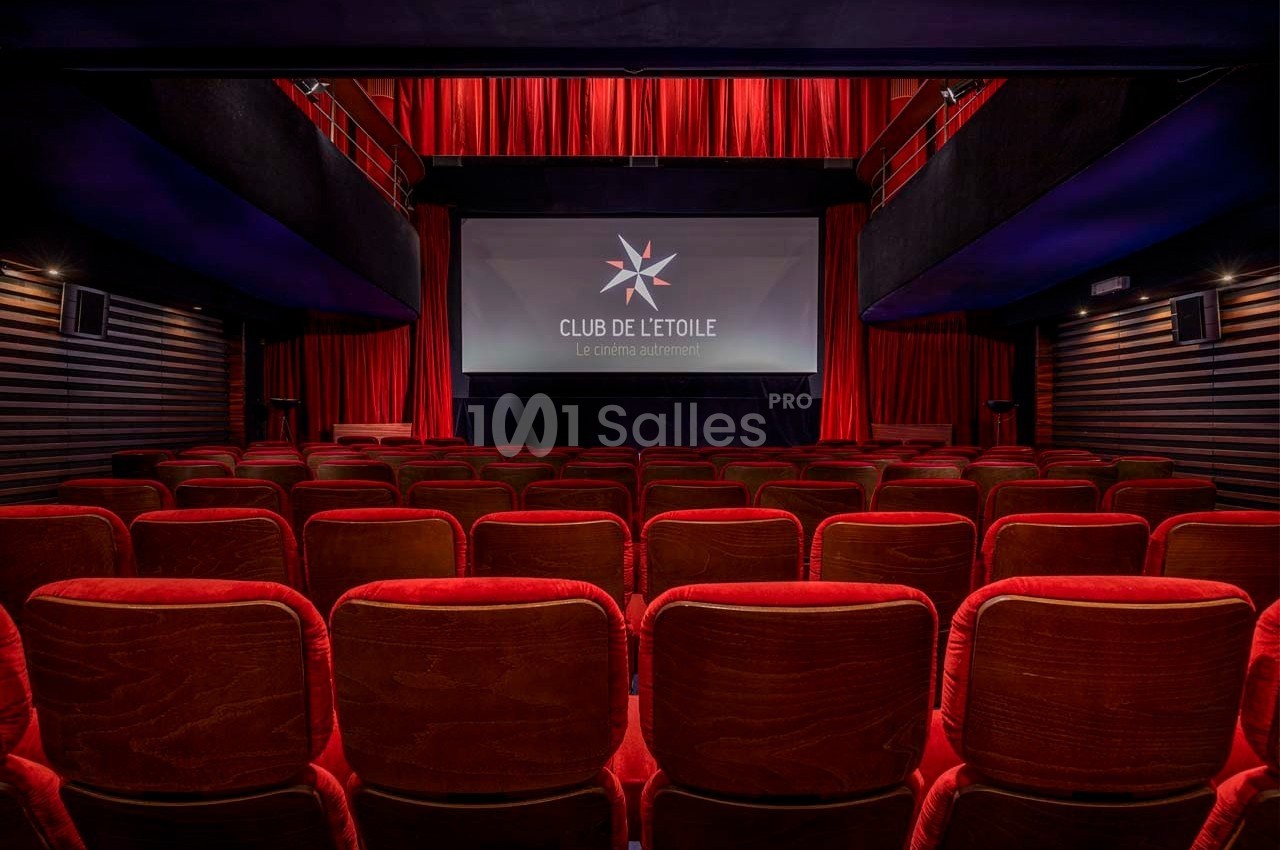 Salle de cinéma vide avec des fauteuils rouges, un écran allumé affichant un logo et des rideaux rouges en arrière-plan.
