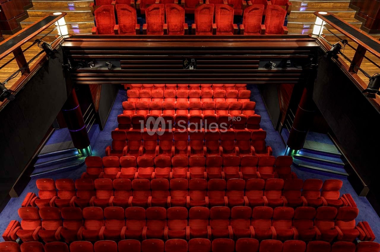 Salle de théâtre avec plusieurs rangées de sièges rouges vides et un balcon supérieur en bois.