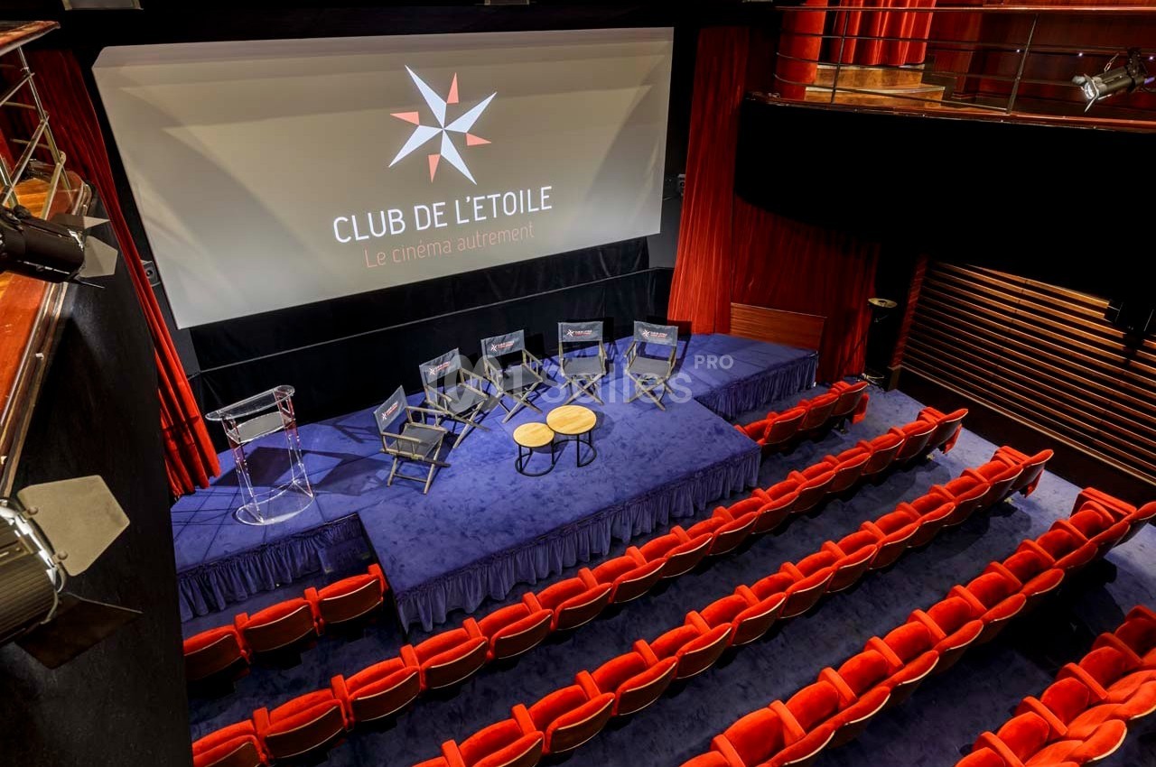 Salle de cinéma avec écran, estrade équipée de chaises et table ronde, entourée de sièges rouges.