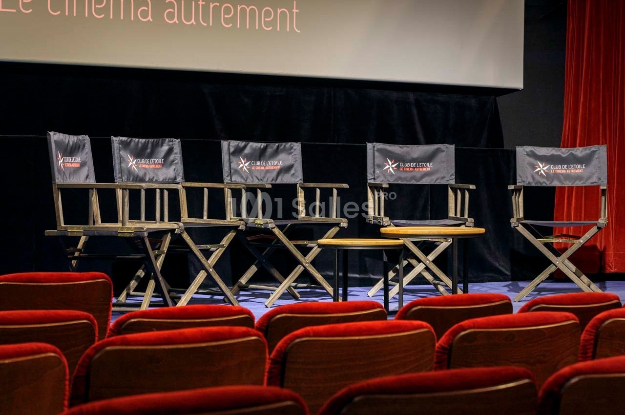 Rangée de fauteuils de réalisateur vides alignés devant un écran de cinéma dans une salle avec rideaux rouges.