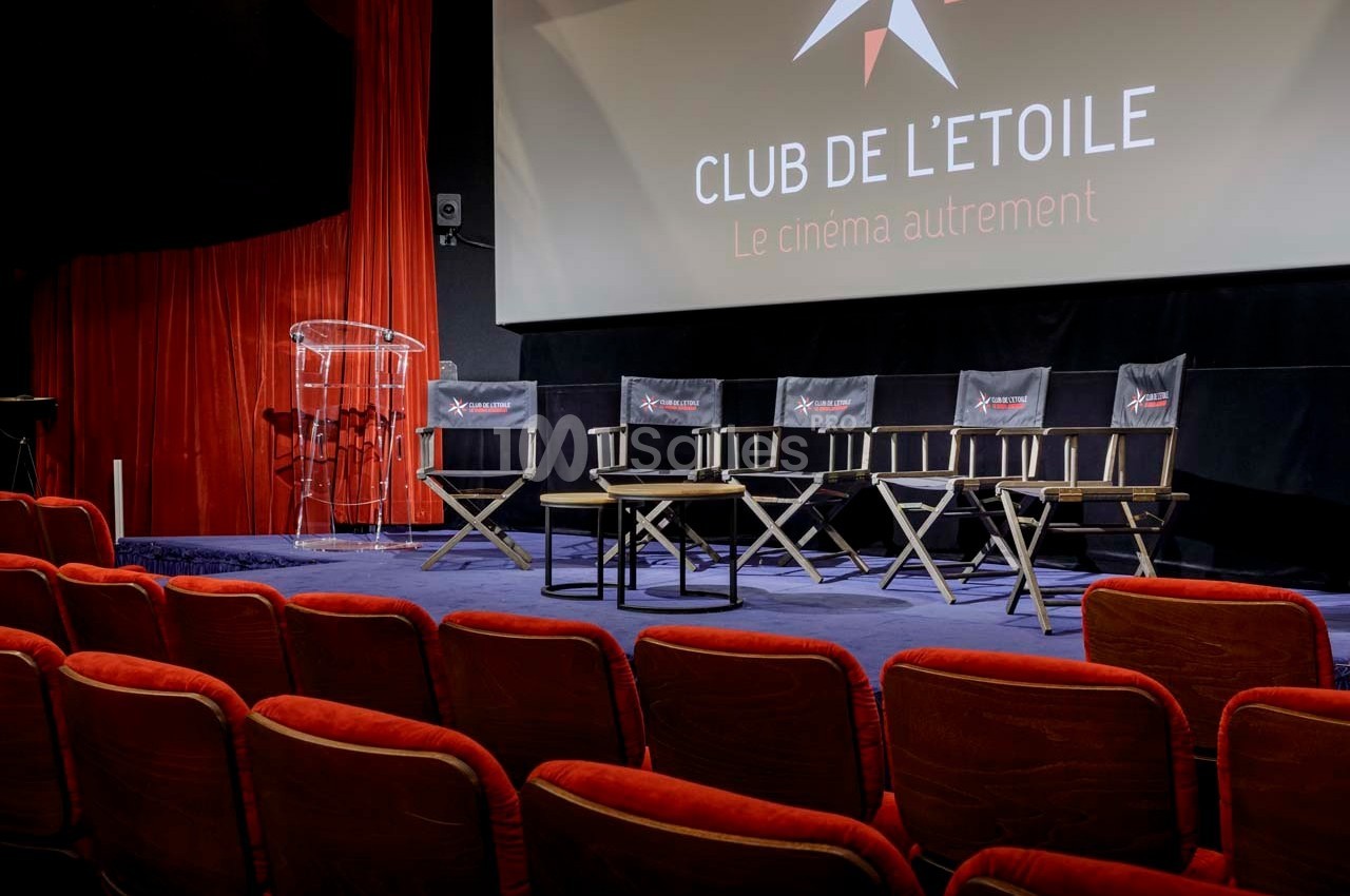 Salle de cinéma avec fauteuils rouges, estrade équipée de chaises pliantes et pupitre transparent devant un écran.