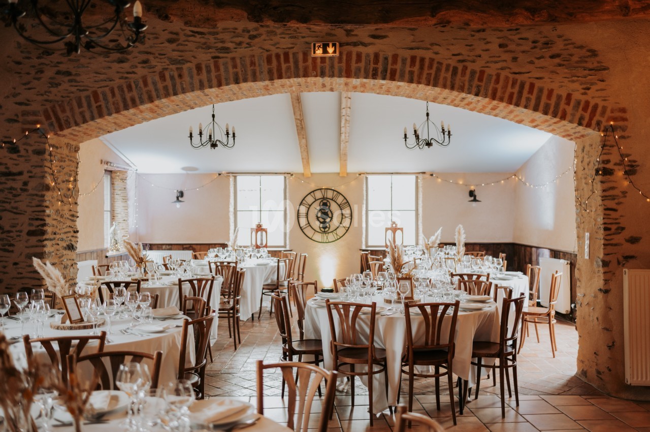 Salle de réception décorée avec des tables rondes dressées, des chaises en bois et des guirlandes lumineuses suspendues.