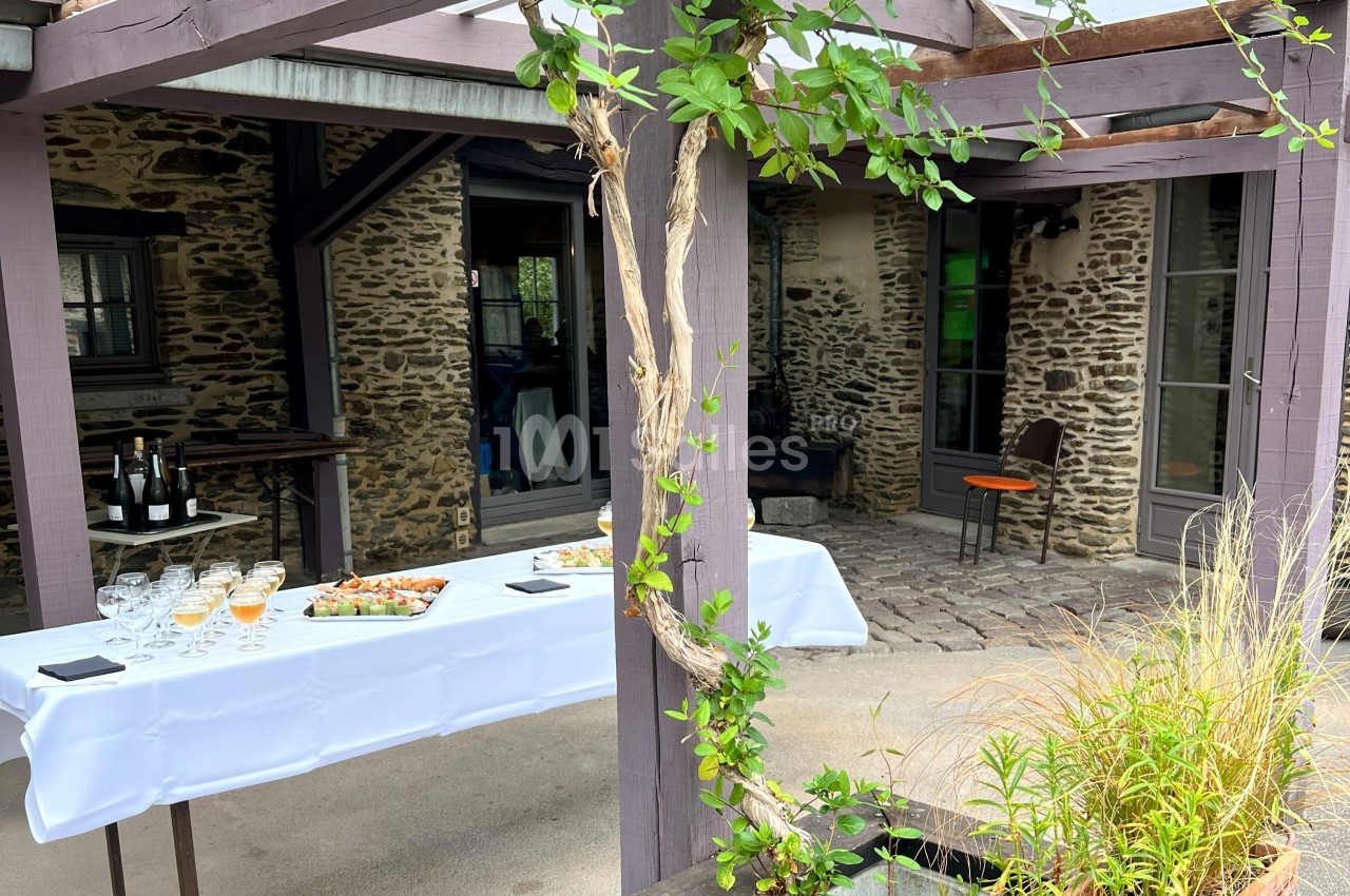 Table dressée avec verres, bouteilles et plateaux de nourriture sous une pergola en bois, devant un bâtiment en pierre.