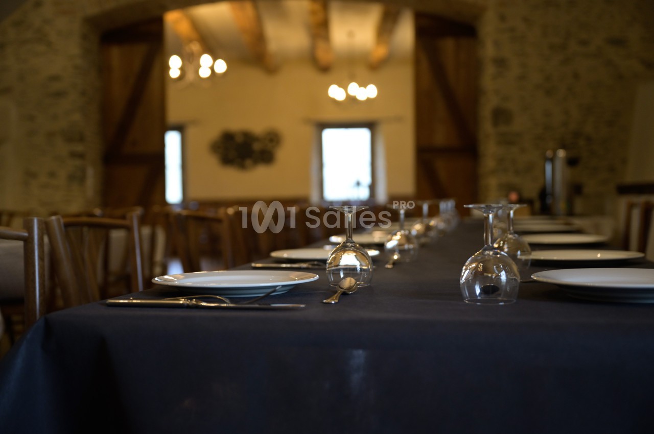 Table dressée avec assiettes, couverts et verres dans une salle en pierre avec poutres apparentes et éclairage chaleureux.