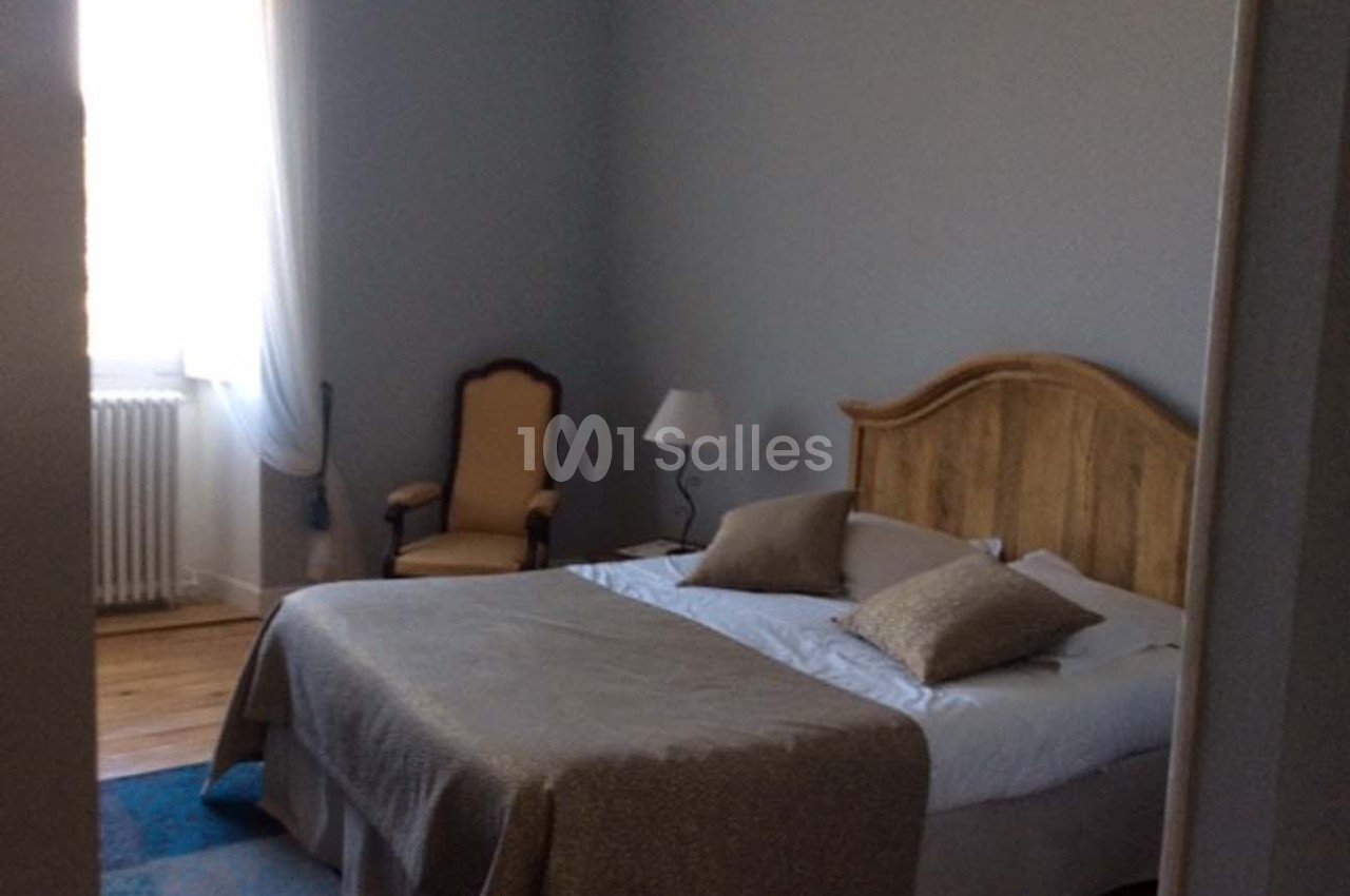 Chambre avec lit double, tête de lit en bois, fauteuil, lampe de chevet et lumière naturelle provenant d'une fenêtre.