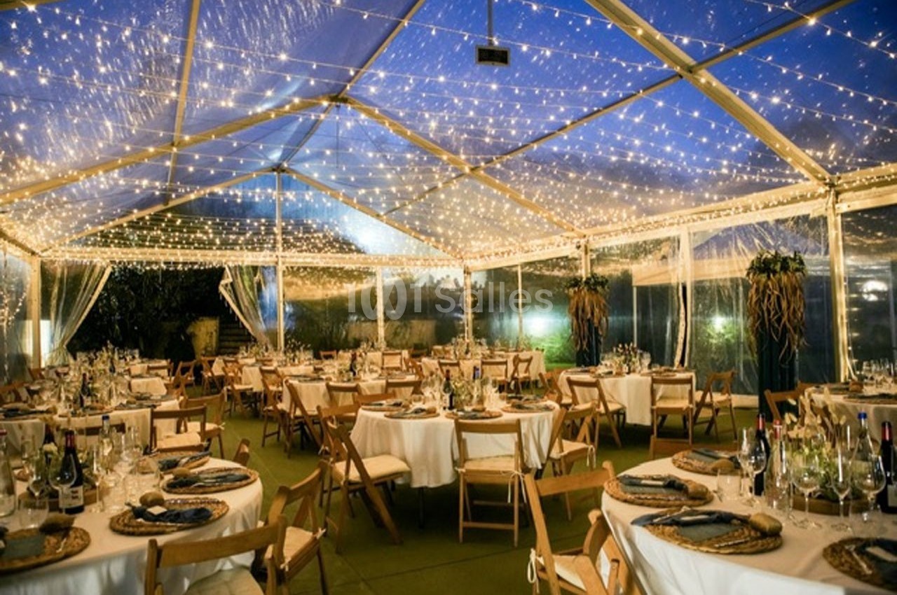 Salle de réception sous une tente transparente illuminée par des guirlandes, avec tables dressées pour un événement.