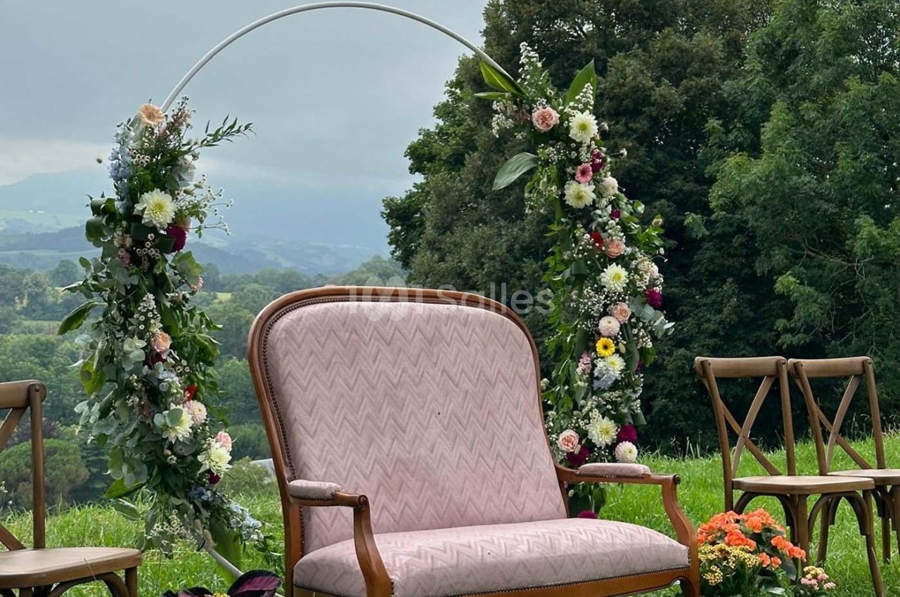 Arche florale décorée avec des fleurs colorées, fauteuil rose et chaises en bois dans un cadre naturel verdoyant.