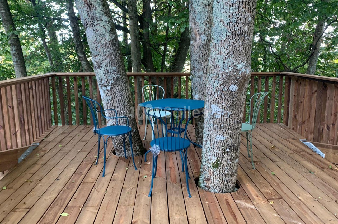Terrasse en bois entourant des arbres, avec des chaises et une table en métal bleu disposées au centre.