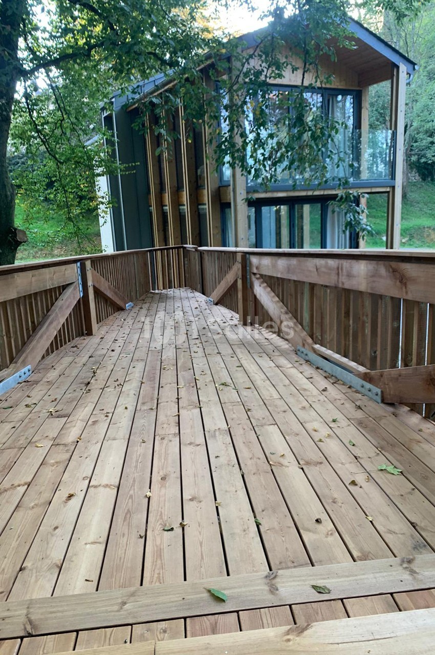 Passerelle en bois menant à une maison moderne entourée d'arbres dans un environnement naturel.