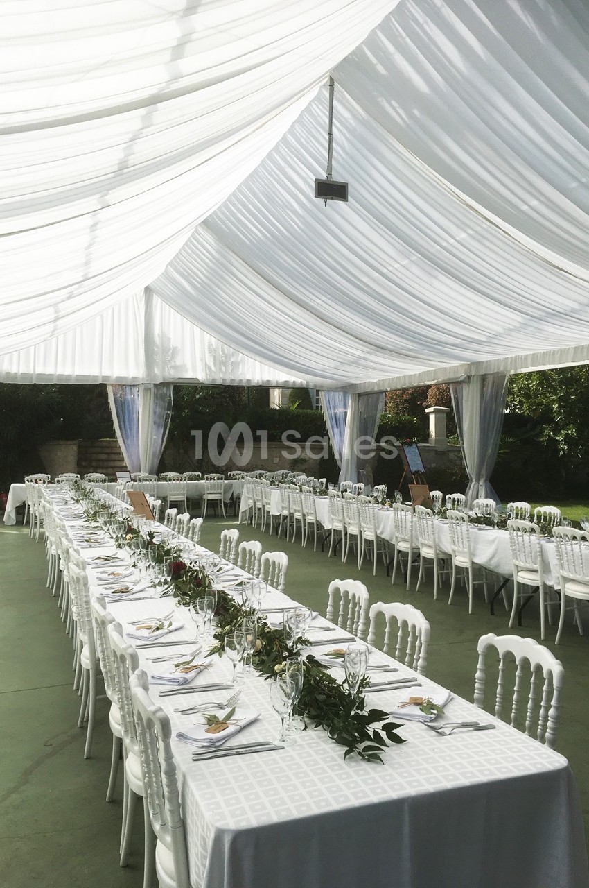 Salle de réception extérieure avec tables alignées, nappes blanches, chaises assorties et décoration végétale sous une tente.