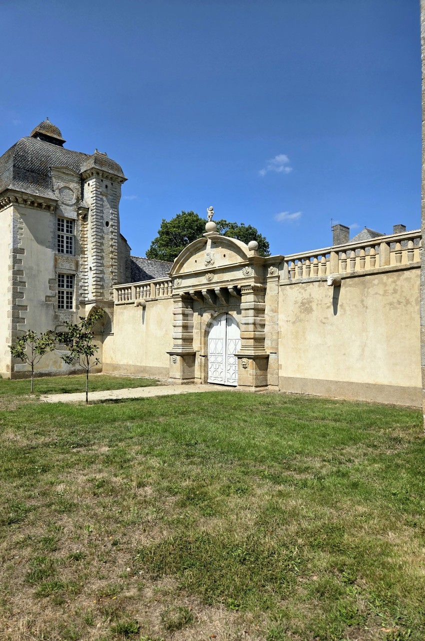 Façade d'un bâtiment historique avec une porte ornée, entourée d'un mur en pierre et d'une pelouse.