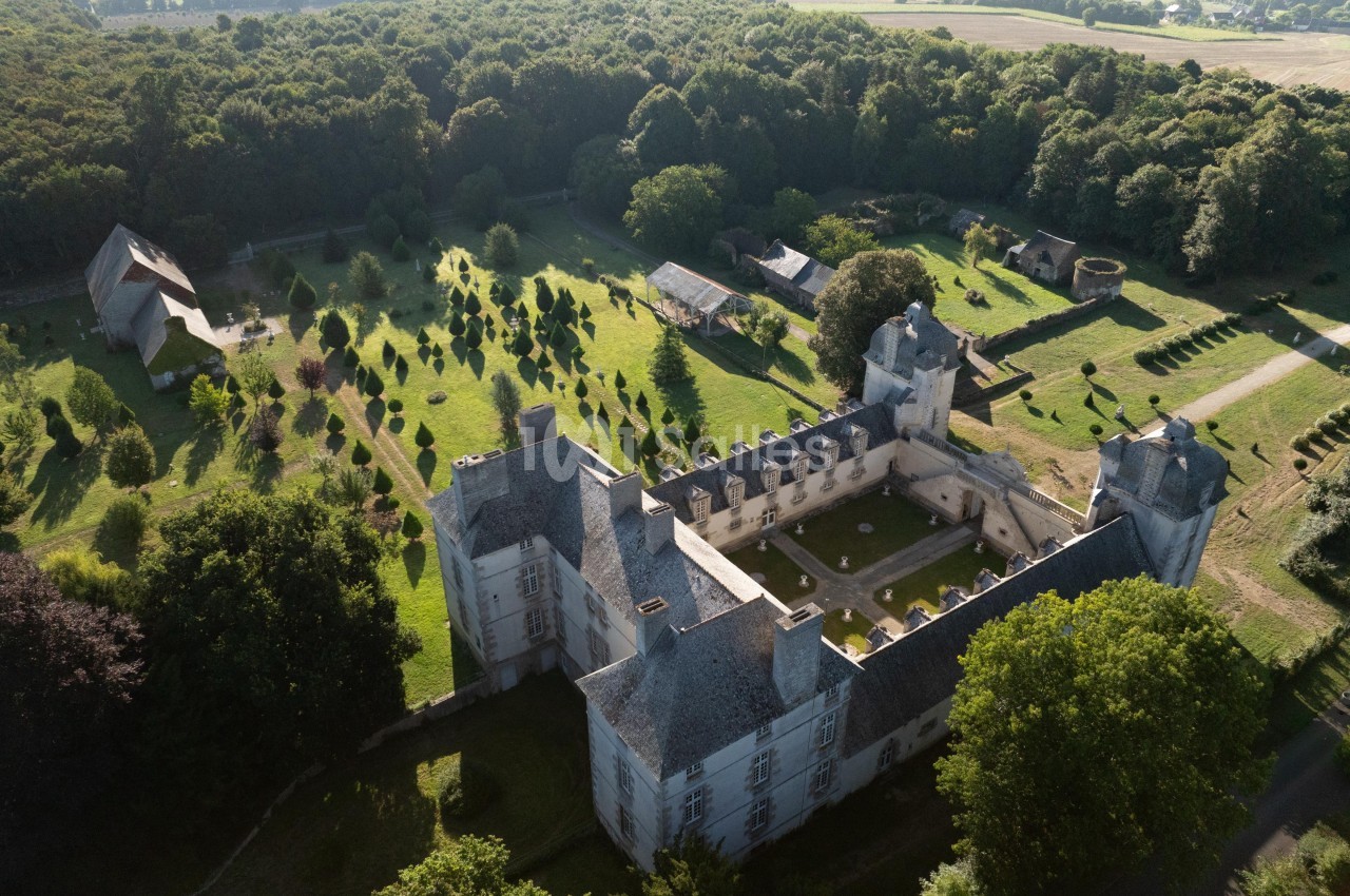 Vue aérienne d'un château entouré de jardins, d'arbres et de bâtiments annexes, au cœur d'une campagne verdoyante.