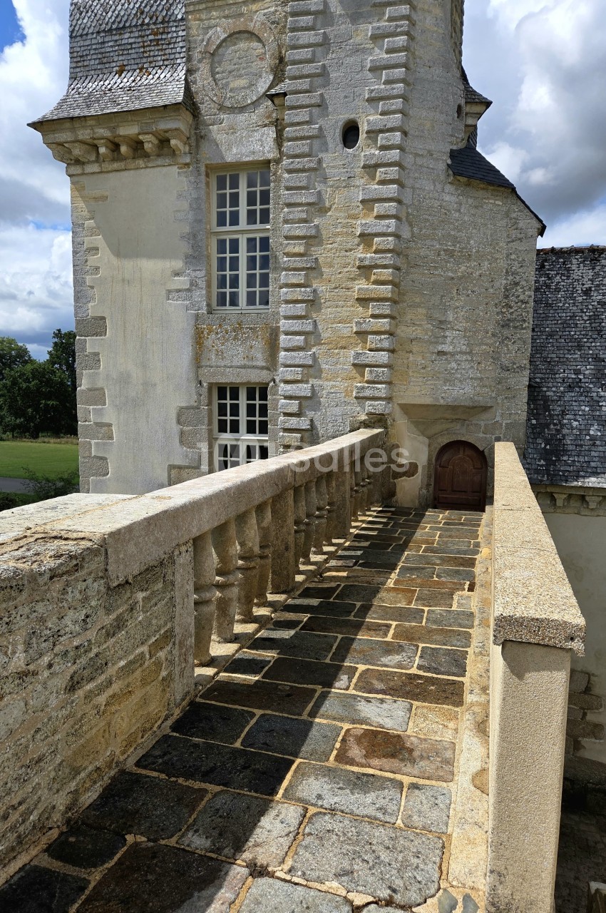 Passerelle en pierre bordée de balustrades menant à une porte en bois sur une tour en pierre ancienne.