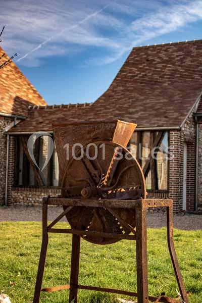 Machine agricole ancienne en métal rouillé, posée sur une pelouse devant des bâtiments en briques et bois.