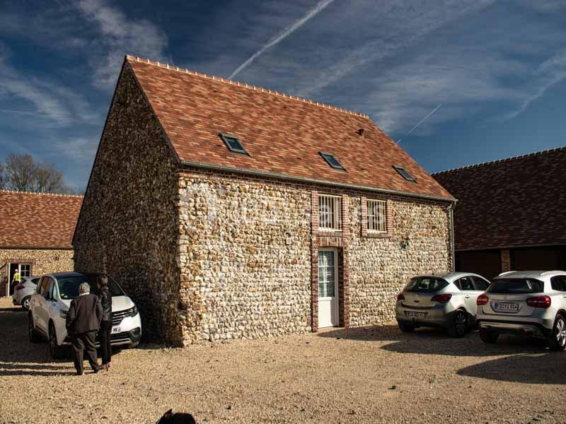 Bâtiment en pierre avec toit en tuiles, entouré de voitures stationnées sur une cour en gravier.