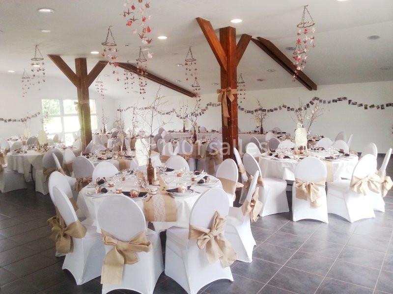Salle décorée pour un événement avec des tables rondes dressées, nappes blanches et chaises ornées de nœuds beiges.