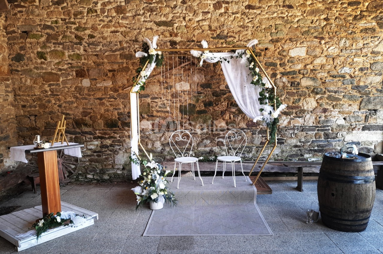 Décor de cérémonie avec arche hexagonale ornée de fleurs blanches, deux chaises et un pupitre en bois devant un mur en…