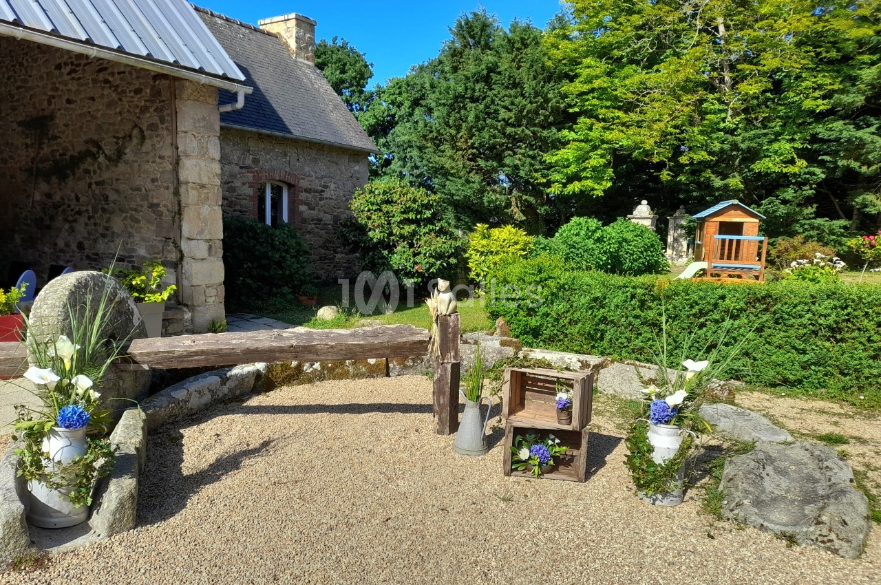 Cour extérieure avec décor champêtre, comprenant des fleurs, des caisses en bois et une petite maison en arrière-plan.