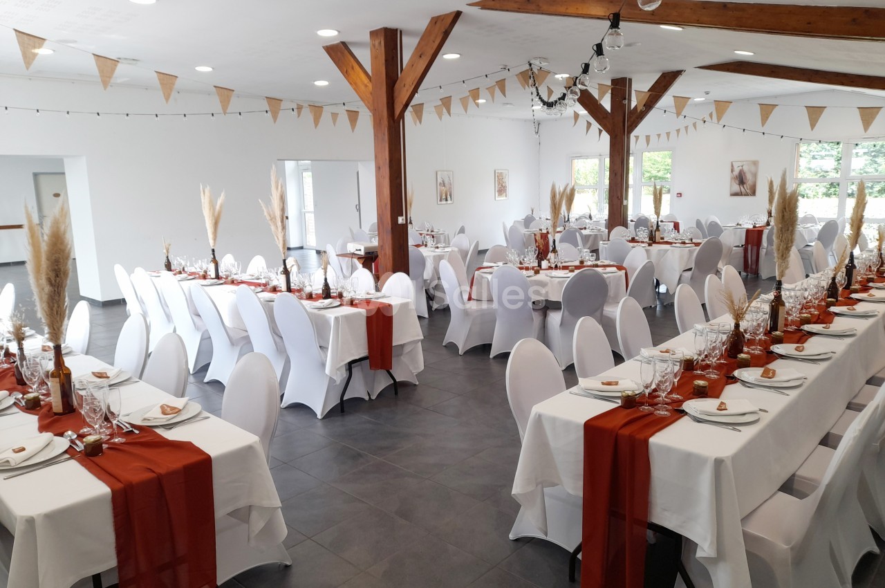 Salle de réception décorée pour un événement, avec tables dressées, nappes blanches et chemins de table rouges.