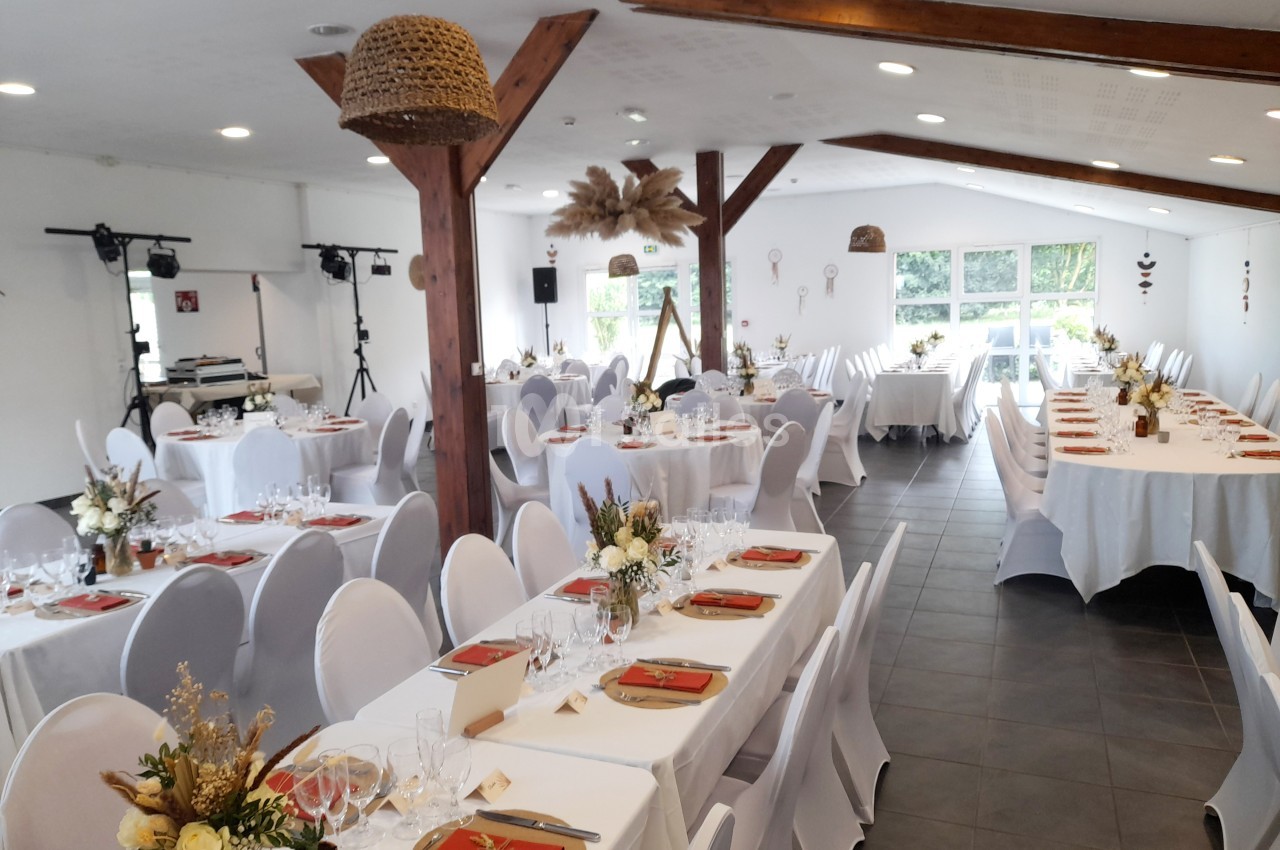 Salle de réception décorée avec des tables dressées, nappes blanches, chaises houssées et arrangements floraux.