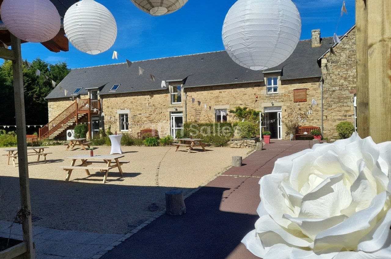 Cour d'une bâtisse en pierre avec lanternes blanches suspendues, tables en bois et grande rose blanche au premier plan.