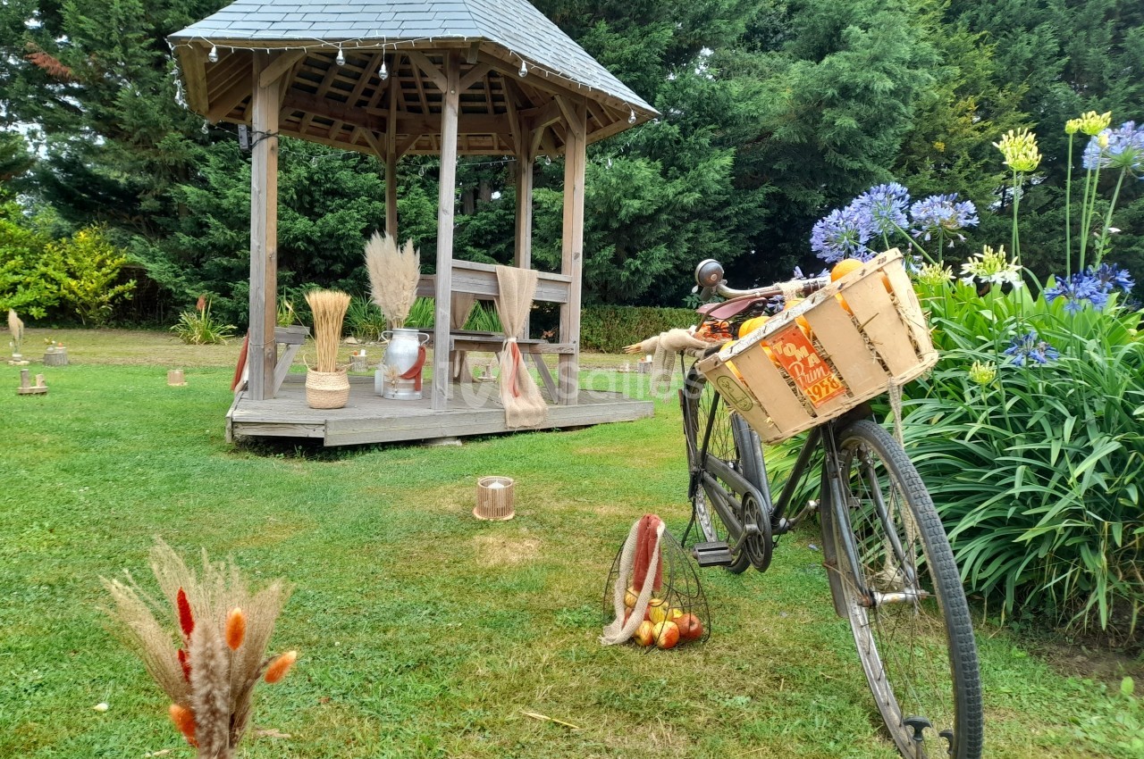 Un vélo ancien avec un panier rempli de pommes est posé près d'un kiosque en bois dans un jardin verdoyant.