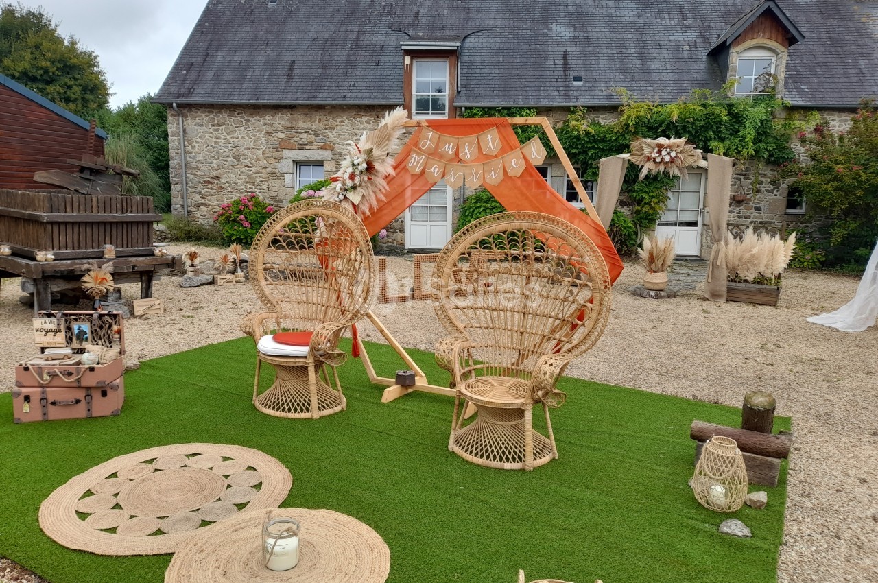 Espace extérieur décoré pour un événement avec fauteuils en rotin, arche orange et éléments bohèmes devant une maison en…