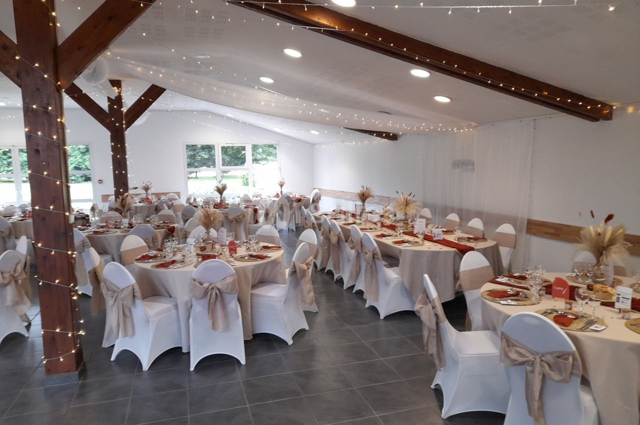 Salle de réception décorée avec des tables rondes, nappes blanches, chaises habillées et guirlandes lumineuses suspendues.