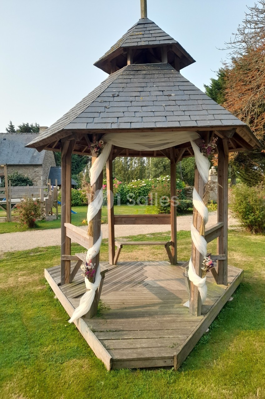 Kiosque en bois décoré de voiles blancs et de fleurs, situé sur une pelouse dans un jardin.