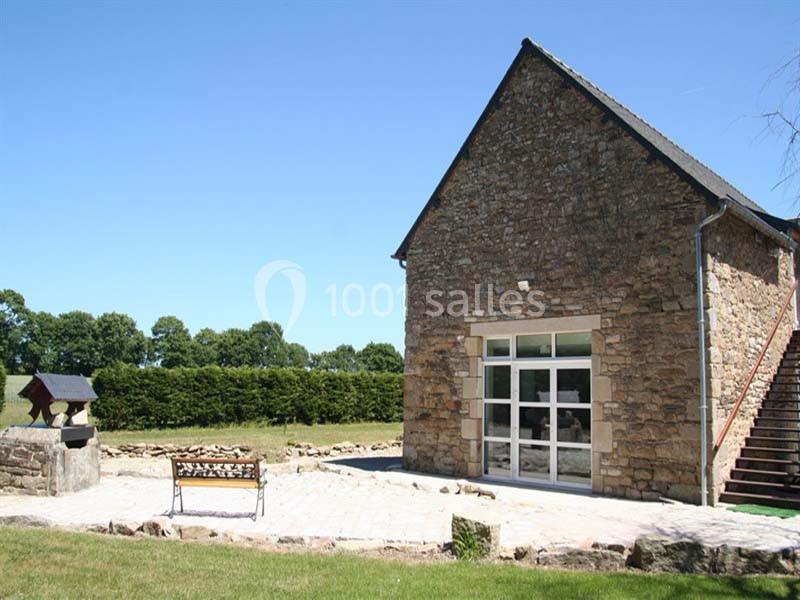 Façade d'une maison en pierre avec une grande baie vitrée, entourée d'une cour pavée et d'un jardin verdoyant.