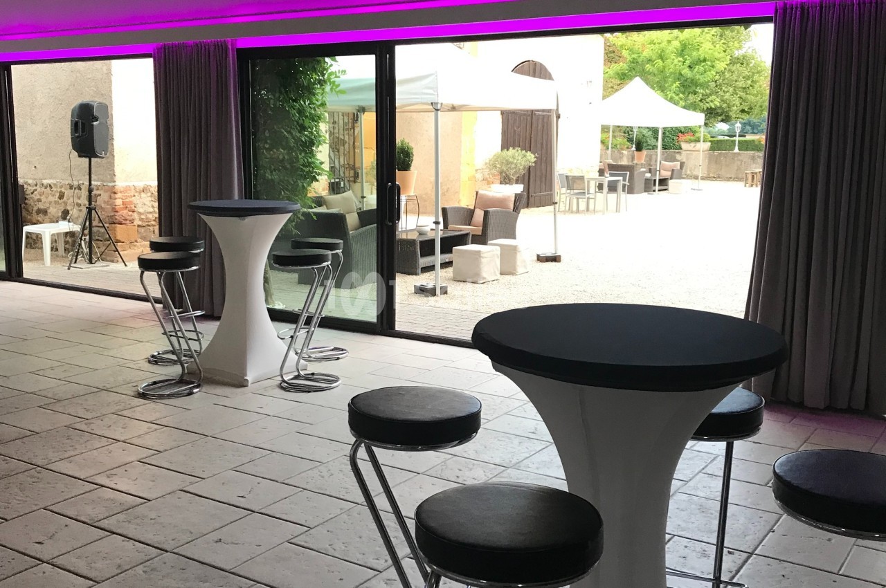 Salle avec tables hautes et tabourets, donnant sur une terrasse extérieure aménagée visible à travers de grandes baies…