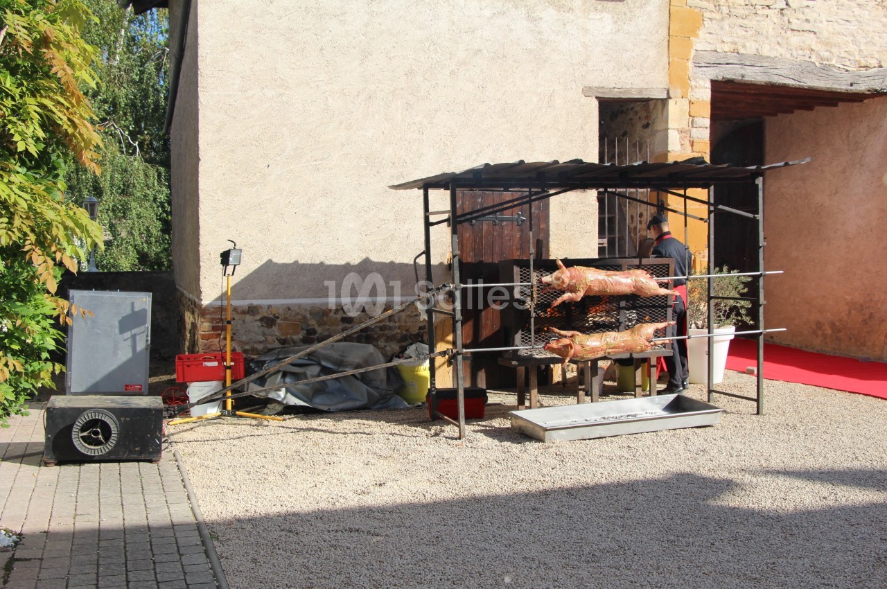 Deux cochons rôtissent sur une broche devant un bâtiment en pierre, dans une cour ensoleillée.