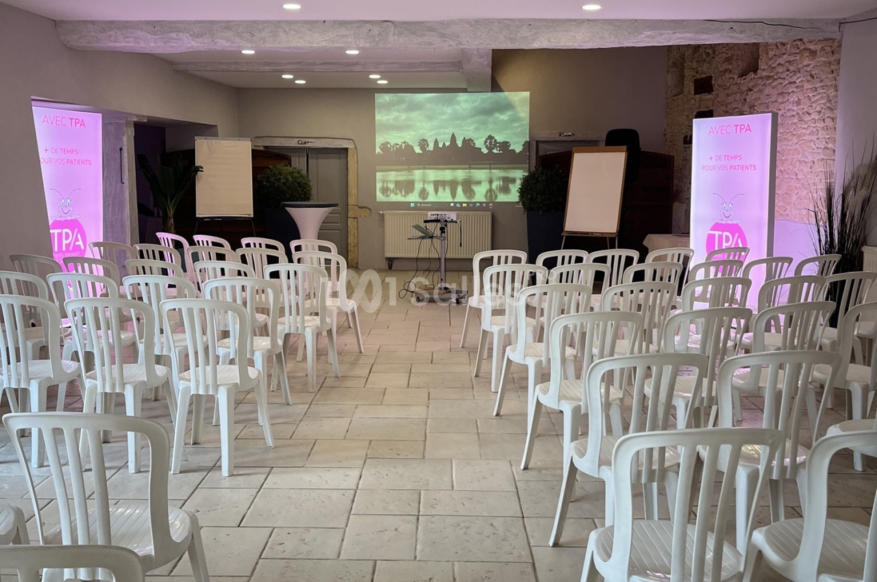 Salle de conférence avec des rangées de chaises blanches, un écran de projection allumé et des affiches promotionnelles.