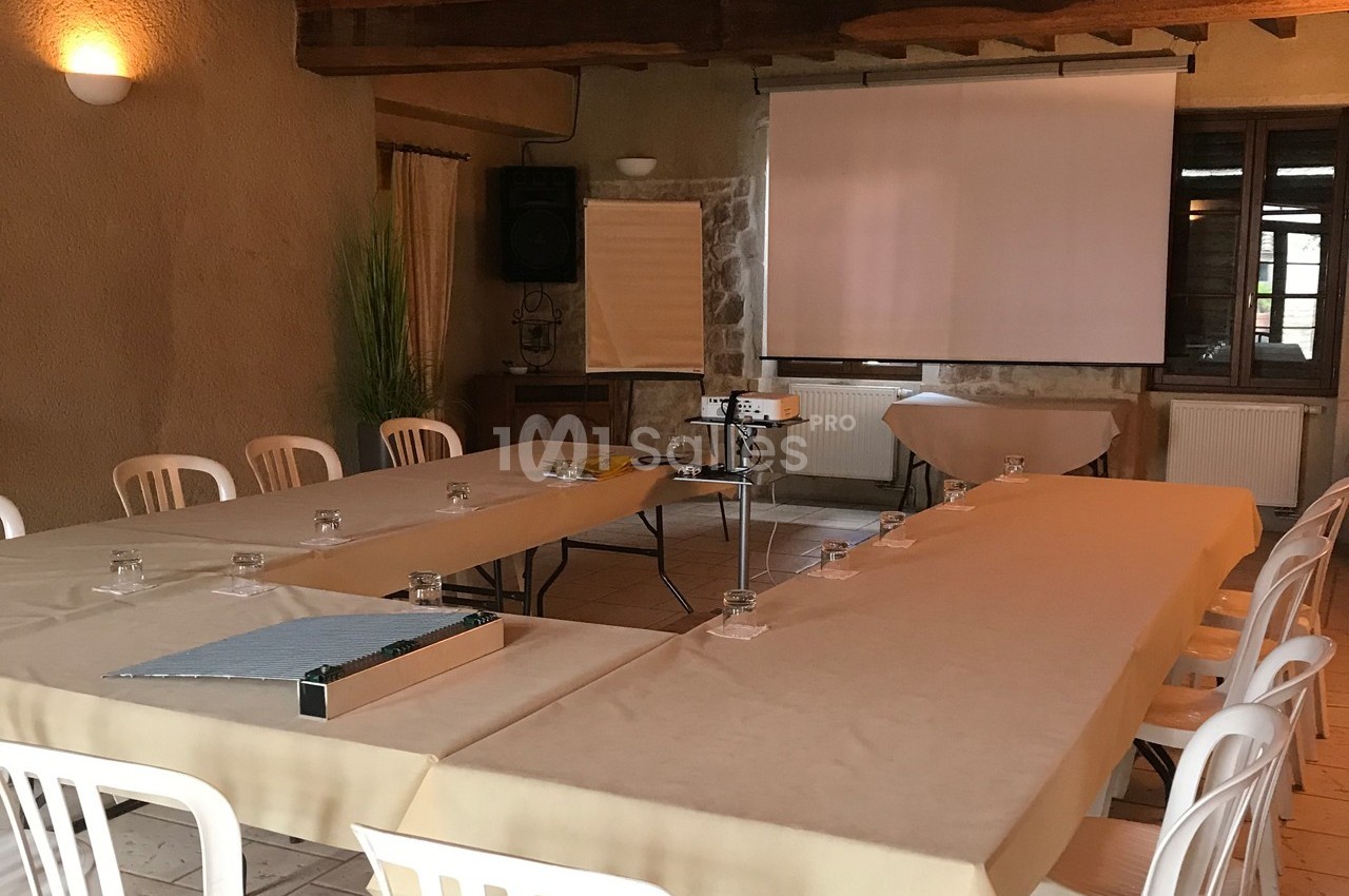 Salle de réunion avec tables disposées en U, chaises blanches, projecteur, écran et tableau de présentation.