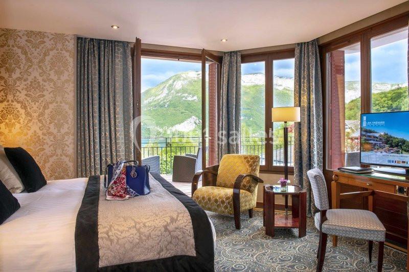 Chambre d'hôtel lumineuse avec lit double, fauteuils, bureau, télévision et balcon offrant une vue sur des montagnes…
