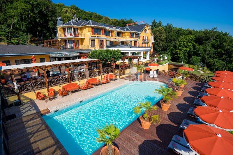 Hôtel avec piscine extérieure entourée de transats, parasols rouges et végétation verdoyante en arrière-plan.