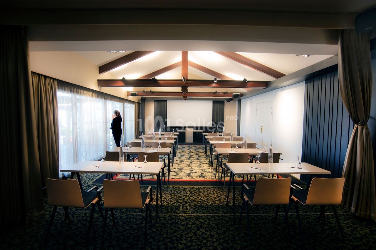 Salle de conférence lumineuse avec des rangées de tables et de chaises, éclairée par des fenêtres et des poutres apparentes.