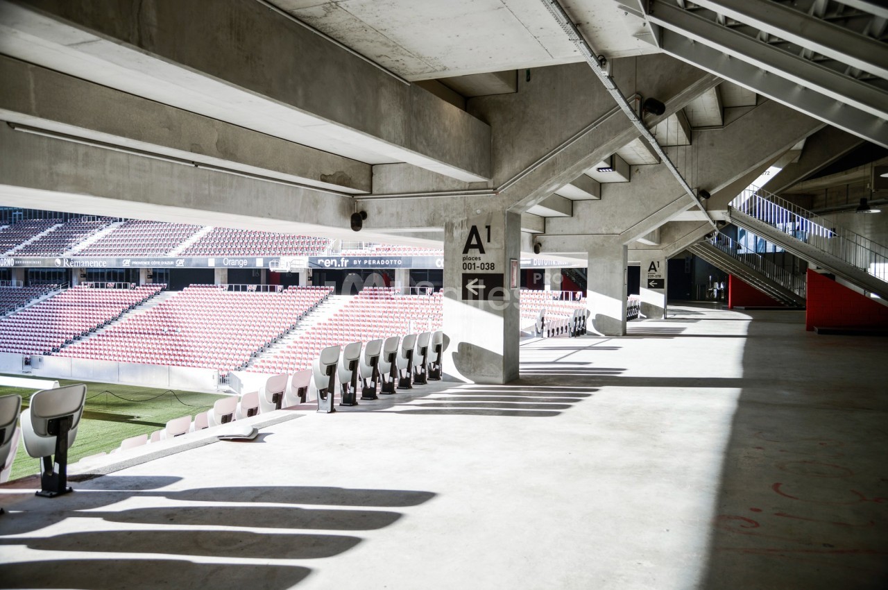 Vue intérieure d'un stade vide montrant des gradins, des sièges et une structure en béton sous un toit partiellement ouvert.