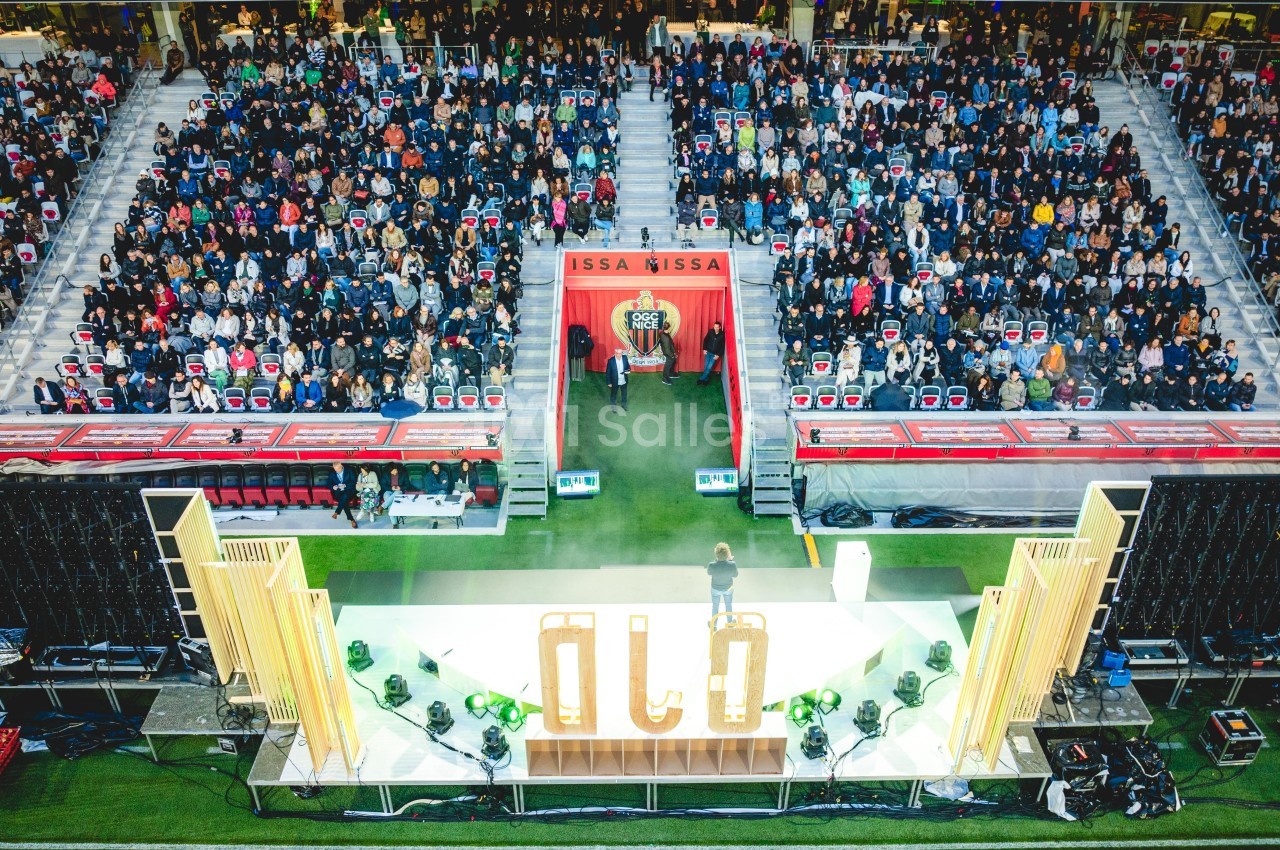 Vue d'une scène éclairée dans un stade rempli de spectateurs, avec des gradins et une décoration lumineuse.