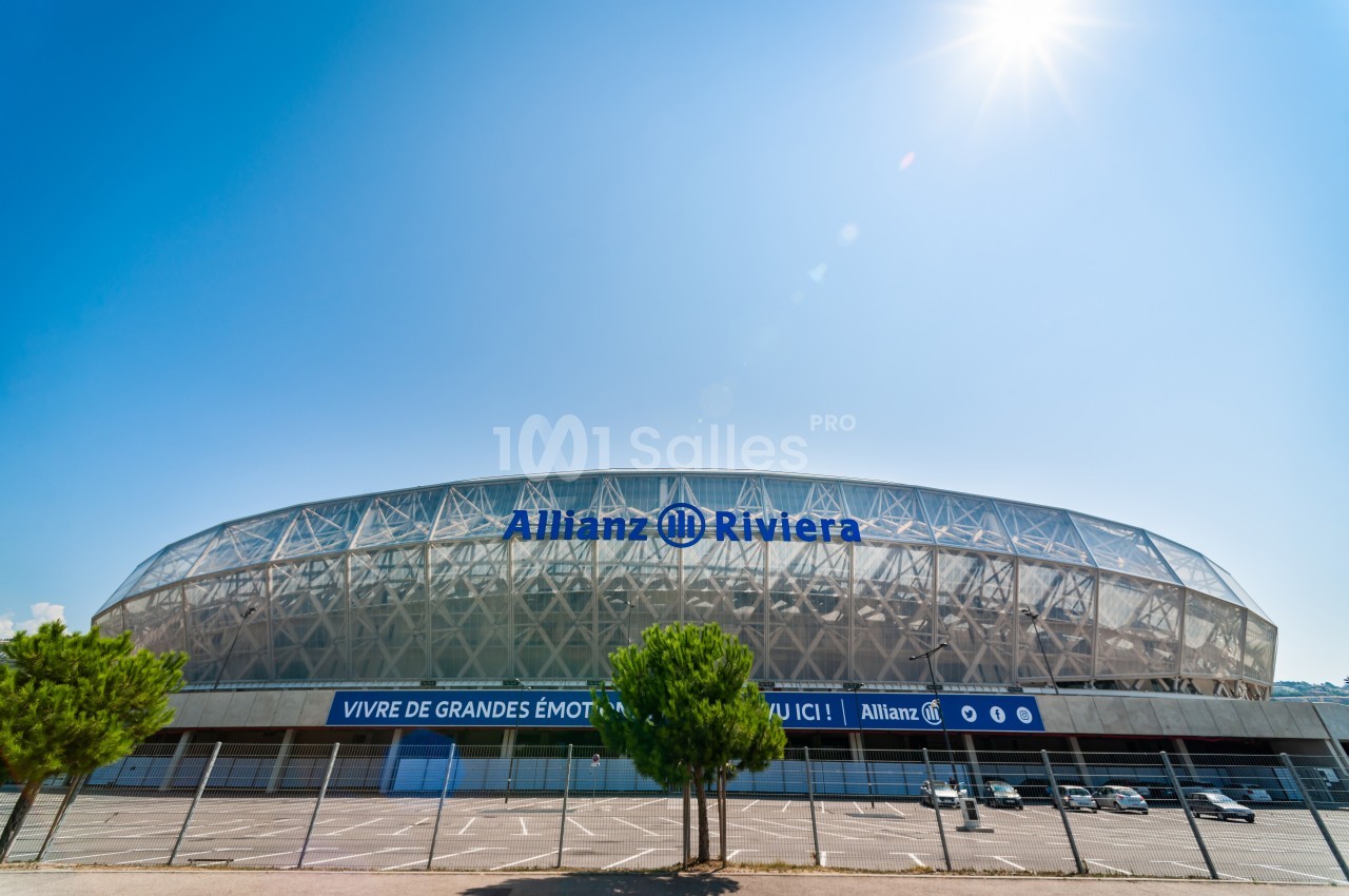 Stade Allianz Riviera sous un ciel ensoleillé, entouré de quelques arbres et d'un parking vide au premier plan.