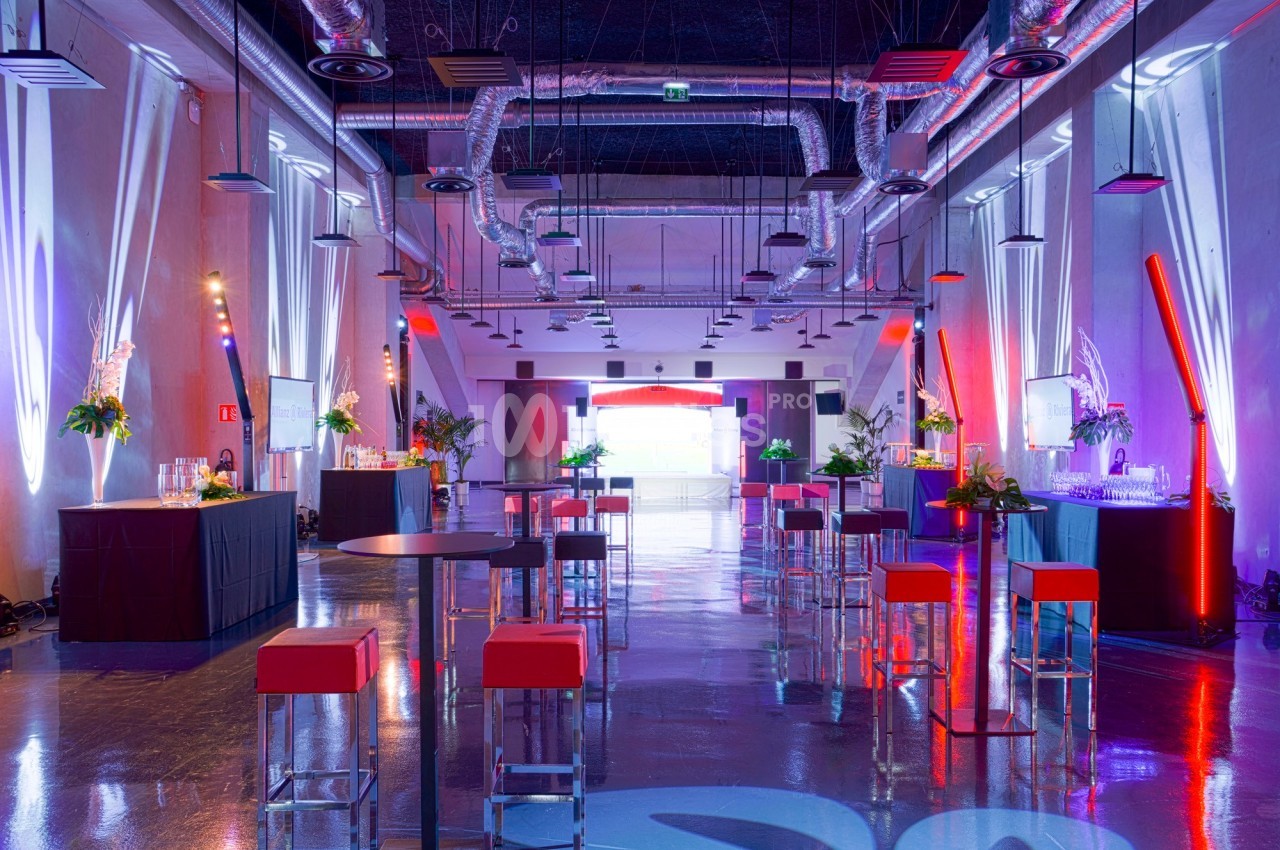 Salle moderne aménagée pour un événement, avec tables hautes, tabourets rouges et éclairage coloré.