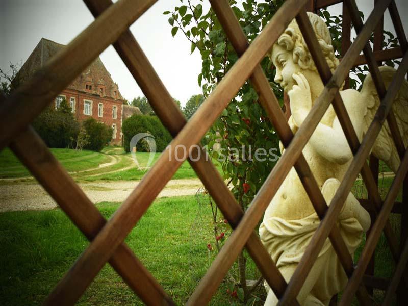 Statue d'ange en pierre vue à travers une clôture en bois, avec une maison en briques rouges en arrière-plan.