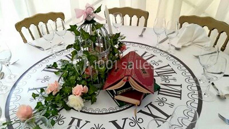 Centre de table décoratif avec livres ouverts, roses, lierre et cage ornée d'un ruban rose sur une nappe à motif d'horloge.