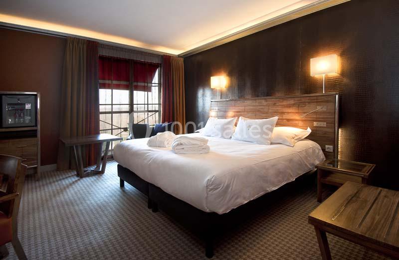 Chambre d'hôtel avec lit double, éclairage chaleureux, table, chaises, et fenêtre avec rideaux rouges.