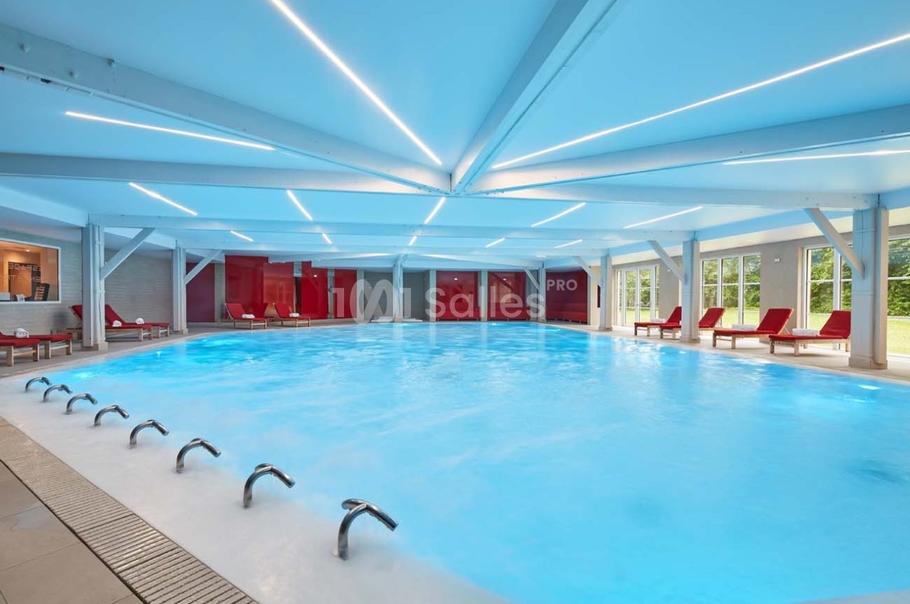 Piscine intérieure lumineuse avec jets d'eau, entourée de chaises longues rouges et grandes baies vitrées.